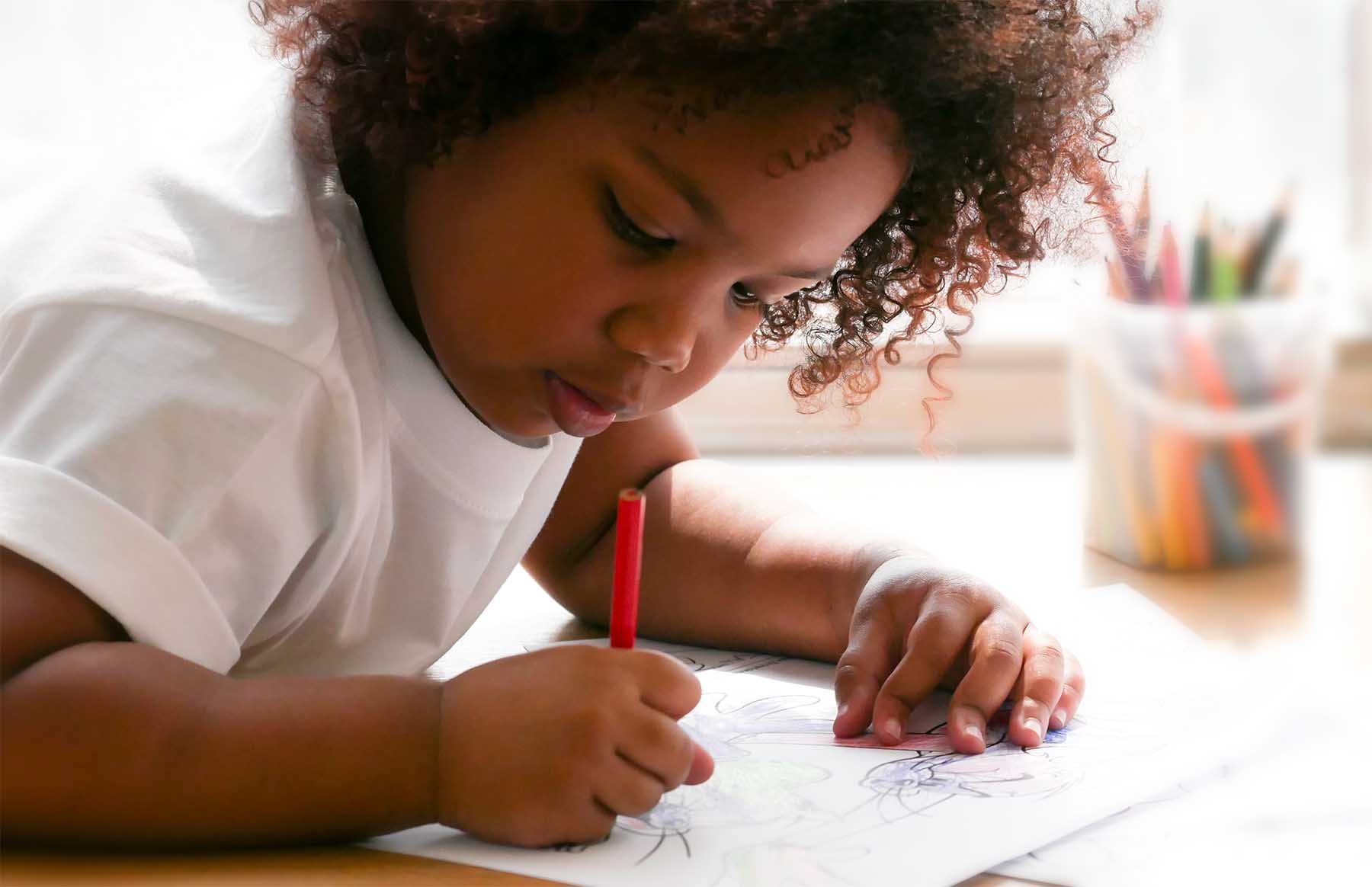 A young girl in a white shirt uses a red pencil to draw on a piece of paper