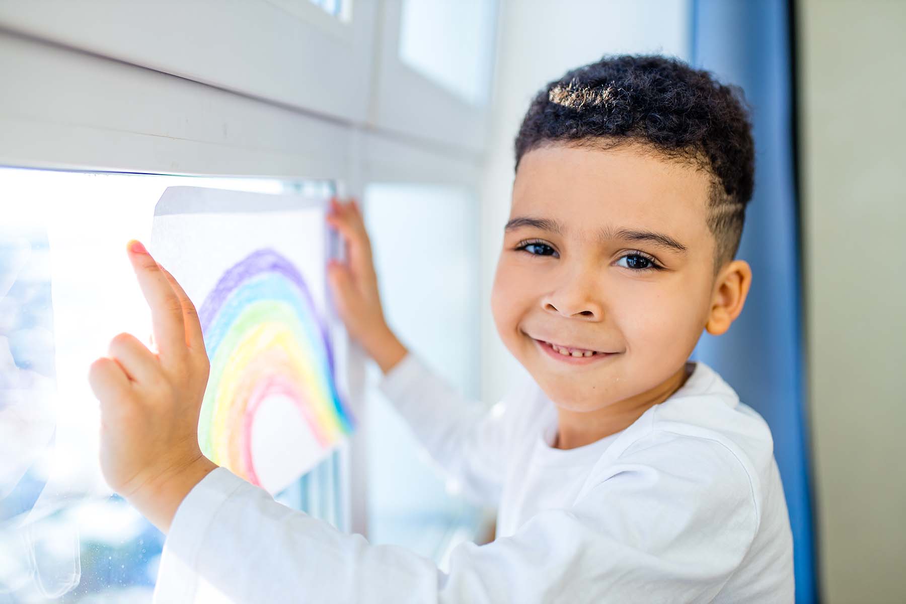 A young child holds his rainbow drawing to the window as he smiles