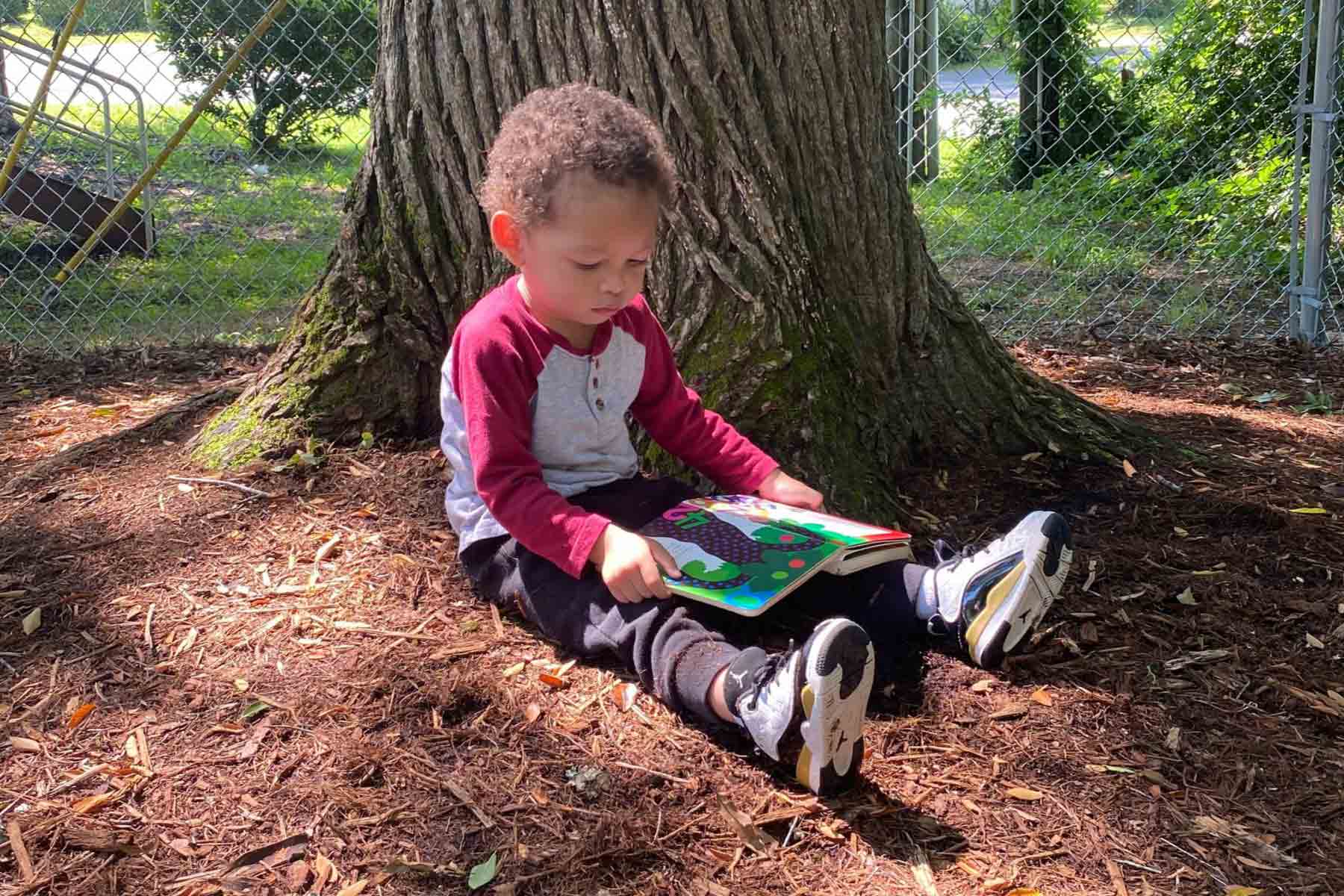 A child in a red and white shirt reading a book next to a tree on mulch at Community School for People Under Six in Carrboro, NC